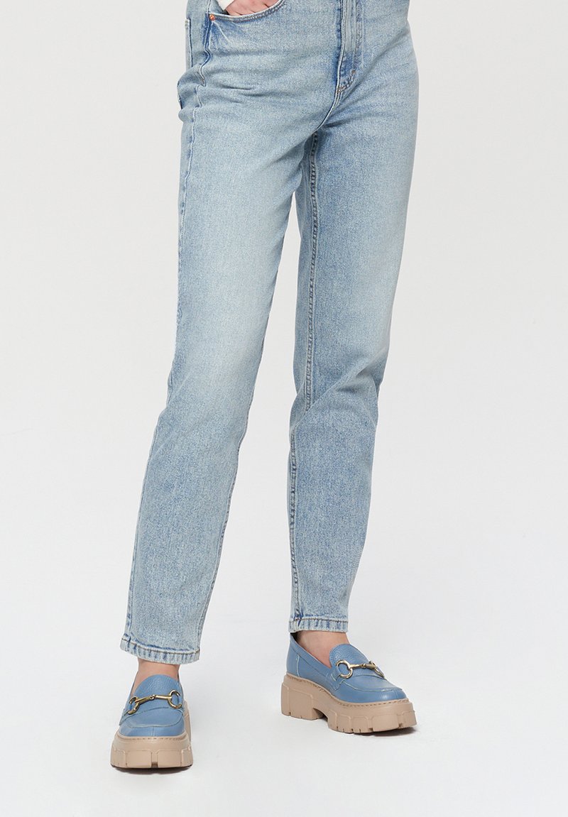 Niebieskie loafersy z masywną beżową podeszwą, z ozdobami w kolorze złota, zestawione z jasnoniebieskimi dżinsami o prostym kroju, z kieszeniami.