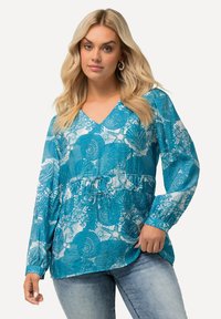 Blusa teal con motivi floreali e circolari bianchi, maniche lunghe e scollo a V. Presenta una vita arricciata e un tessuto morbido e testurizzato.