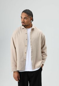 Beige overhemd met knopen, gemaakt van gestructureerde stof, voorzien van een kraag en een losse pasvorm, gedragen over een wit T-shirt. Zwarte broek.