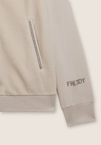Felpa beige con finitura testurizzata, caratterizzata da una tasca con zip ornata da accenti metallici e la scritta "FREDDY" in strass sulla manica.