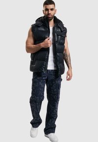 Schwarze Steppweste mit Kapuze, Reißverschluss und gesticktem Markenlogo. Kombiniert mit navy-blauen Camouflage-Cargohosen und weißen Sneakers.