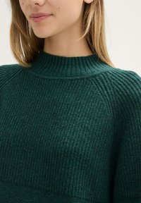 Maglione verde a coste con un alto collo a lupetto. Presenta una superficie strutturata e maniche raglan. Materiale morbido, design leggero.