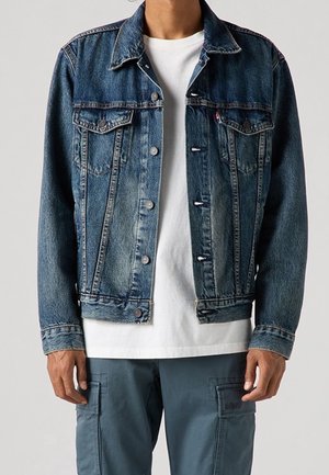 Personne portant une veste en jean bleue par-dessus un t-shirt blanc et un pantalon cargo bleu, debout devant un fond clair uni.