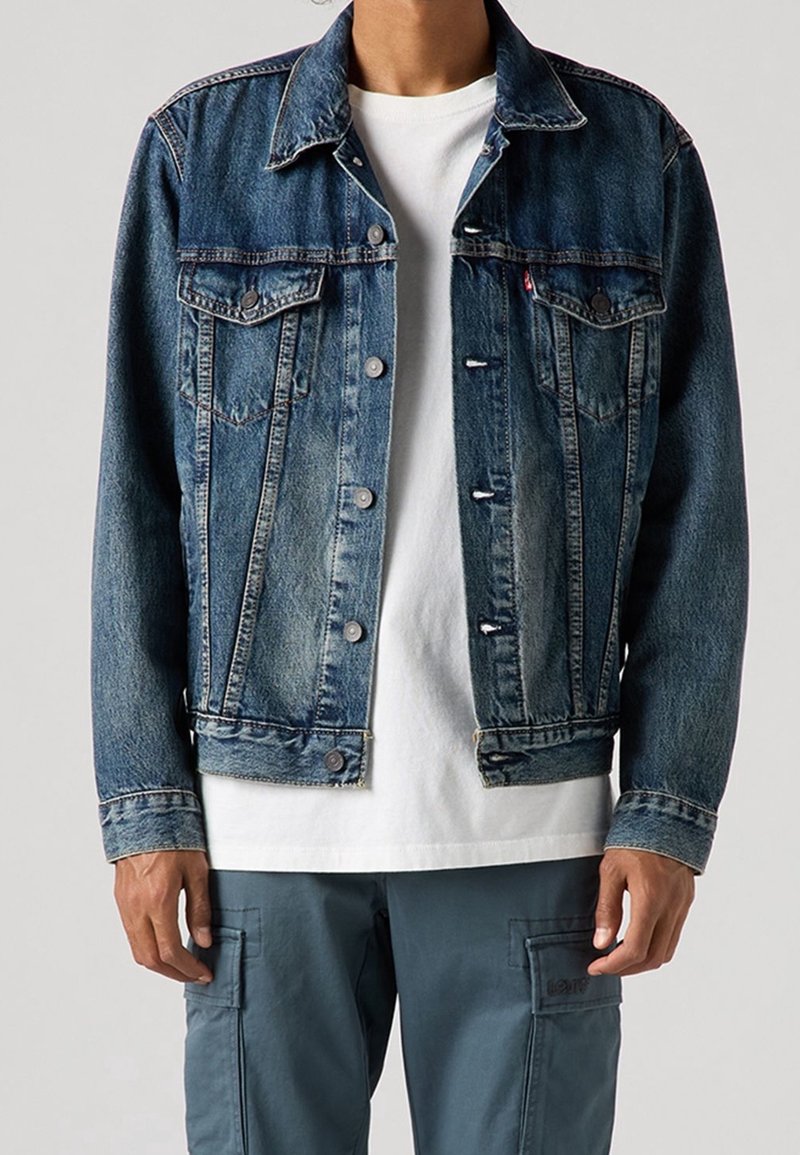 Personne portant une veste en jean bleue par-dessus un t-shirt blanc et un pantalon cargo bleu, debout devant un fond clair uni.