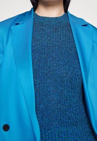 Niebieska marynarka na teksturowanym swetrze o różnorodnych odcieniach niebieskiego i zielonego. Marynarka ma klapy z wycięciem i detal z guzikami.