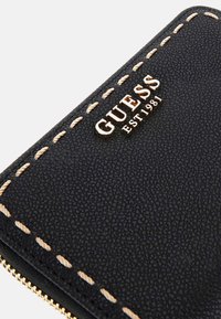 Portefeuille noir en matériau texturé, avec le logo doré "GUESS" et des coutures beige clair le long des bords, fermeture zippée en bas.