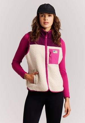 Jonge vrouw met een zwarte pet, een witte en roze fleecebodywarmer over een roze longsleeve, met één hand in de zak van het bodywarmer.