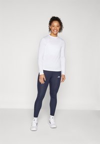 Hvit langermet genser og marineblå tights, begge tettsittende. Funksjoner inkluderer rund halslinje og Adidas-logo på tightsene. Hvite treningssko.