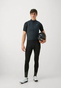 Herren-Radoutfit mit einem dunkelblauen ärmellosen Trikot und engen schwarzen Leggings. Der Radfahrer hält einen schwarzen Helm und trägt weiße Schuhe.
