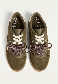 Une paire de baskets en nubuck vert olive avec des lacets à motifs blancs et rouges contrastants, à bout arrondi et semelle en caoutchouc sur fond blanc.