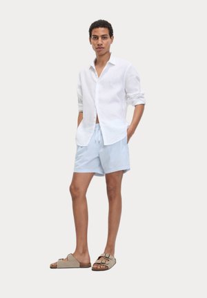 Camicia bianca in lino, pantaloni corto a righe blu chiaro e bianchi, e ciabatte beige. Il modello sta in piedi con le mani in tasca contro uno sfondo neutro.