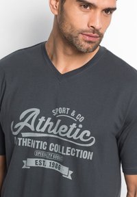 T-shirt gris charbon à col en V en coton, avec un imprimé graphique sportif en gris clair indiquant "Collection Athlétique" dans un design vintage.