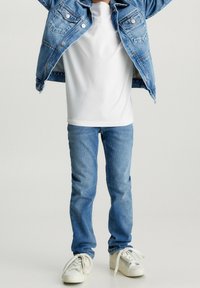 Chaqueta de mezclilla con un tono azul, camiseta blanca debajo, jeans de un azul claro y zapatillas blancas. Conjunto casual con cortes y texturas clásicas.