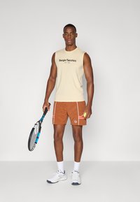 Beige ermeløs skjorte med svart logo, brune shorts med hvite detaljer og rosa striper, holder en tennisracket og en ball, iført hvite joggesko.