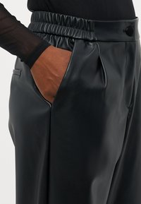 Pantalons en cuir noir avec une taille élastique et des poches latérales. Le tissu a une texture lisse, présentant un léger éclat et des détails plissés.