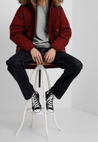 Rode pufferjack met een nep bontcapuchon, grijs T-shirt eronder, donkerblauwe jeans en zwarte high-top sneakers, zittend op een witte kruk.