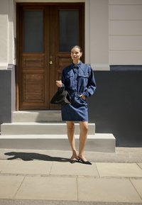 Vrouw in een blauwe denim jurk en zwarte schoenen staat op het voetpad met een zwarte handtas voor een houten deur en een crème wand.