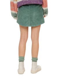 Minifalda de pana verde con dobladillo curvado, combinada con botas blancas hasta el tobillo y calcetines verdes de canalé. Textura suave y diseño ajustado.