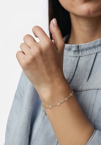 Armbandet i guldton har hjärtformade detaljer och klara ädelstenar, vilande på ett ljusbrunt handled mot en grå-blå topp.