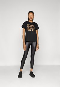 Schwarzes T-Shirt mit einem Leopardenmuster-Graphic und "NY" in Orange, kombiniert mit glänzenden schwarzen Leggings und schwarzen Sneakers.