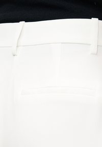 Εγγύτατη λήψη της μέσης ενός λευκού παντελονιού με ραμμένη εφαρμογή, με θηλιές για ζώνη και μία πίσω τσέπη με χαραγή, πάνω σε μαύρο υφασμάτινο φόντο.