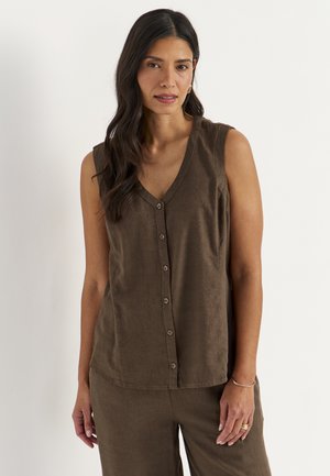 Kvinde med langt mørkt hår iført ærmeløs brun knapbluse og matchende bukser, stående mod en ensfarvet lys baggrund.