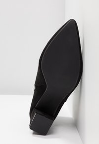 Sort ruskind højhælet sko med spids tå og firkantet hæl. SÅlen er af tekstureret gummi og viser tegn på slid på bunden.