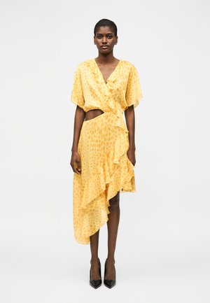 Kvinde iført en gul, asymmetrisk kjole med flæser, udskæring i taljen og korte ærmer, kombineret med sorte spidse hæle.