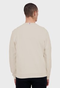 Sudadera beige de manga larga con un escote redondo. Presenta un pequeño detalle de franjas en rojo, blanco y azul en la parte posterior del cuello. Textura suave.