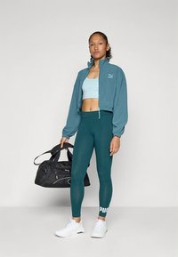 Turkusowa krótka kurtka na jasnoniebieskim topie, do tego ciemnoturkusowe legginsy. Białe akcenty z logo i czarna torba sportowa dopełniają sportowy look.