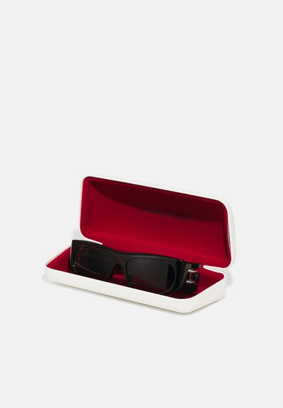 Gafas de sol negras en un estuche duro blanco, forrado con terciopelo rojo. Presentan una forma rectangular y acentos metálicos en los laterales.