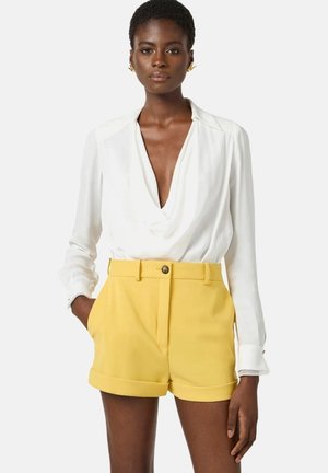 Donna dalla pelle scura che indossa orecchini d'oro, una camicetta bianca drappeggiata e pantaloncini gialli a vita alta con le mani nelle tasche.