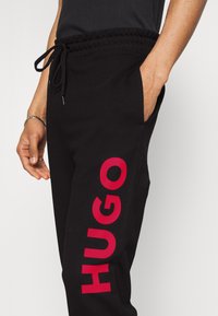 Pantalones de chándal negros con cintura ajustable, que presentan un audaz logo rojo "HUGO" a lo largo del lado izquierdo y un bolsillo en el derecho.