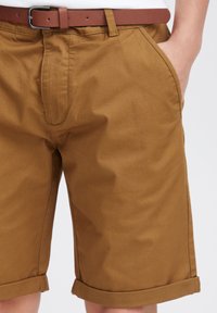 Brown cotton shorts with a smooth texture, featuring rolled cuffs, side pockets, and a brown belt with a metal buckle.