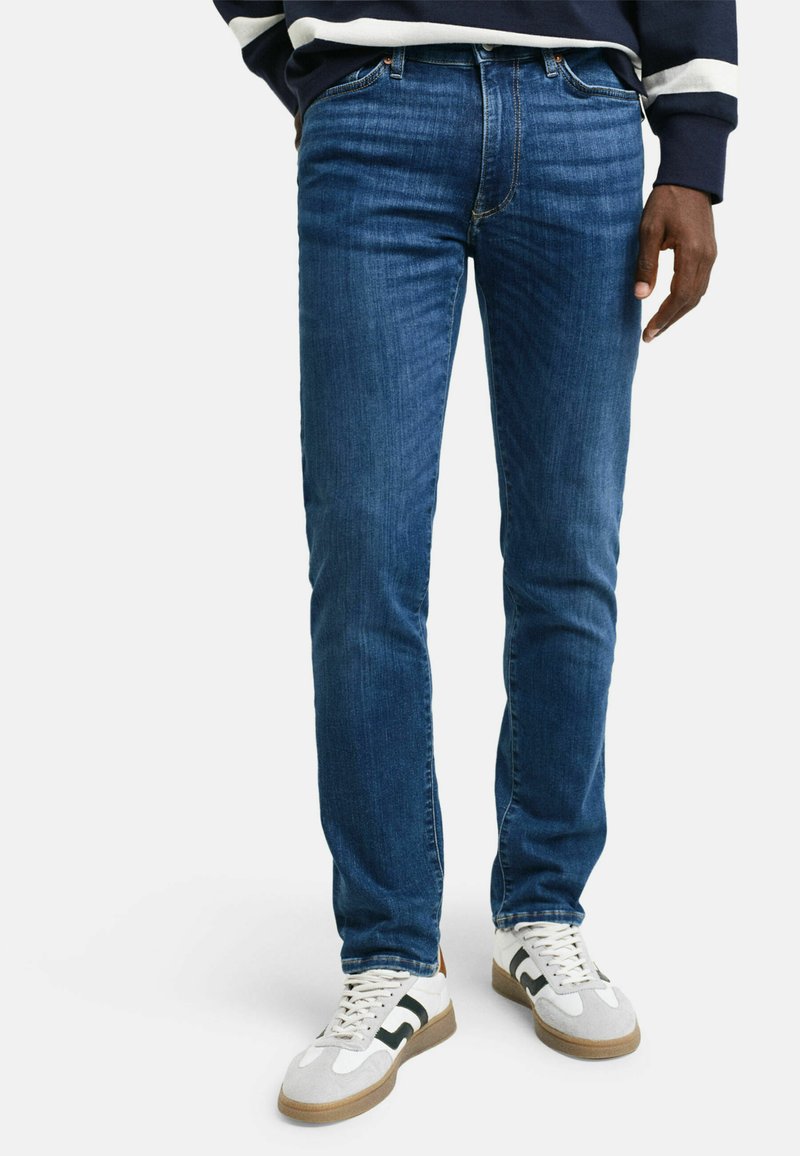 Jeans de mezclilla azul ajustados con un ligero desgaste y diseño de cinco bolsillos, combinados con zapatillas blancas y grises con acentos en verde.