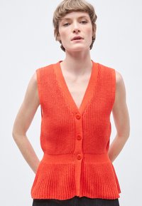 Κόκκινο πλεκτό γιλέκο με V-λαιμόκοψη, κουμπιά στο μπροστινό μέρος και τελείωμα με βολάν. Η υφή του υφάσματος είναι ριγέ, με μονόχρωμο σχέδιο χωρίς μοτίβα.