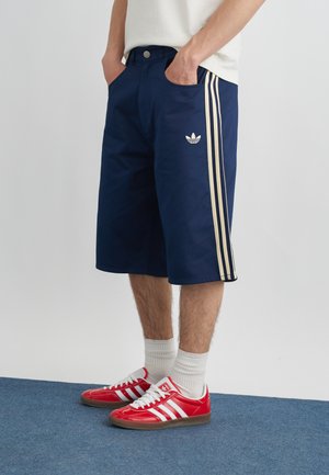 Personne portant un short Adidas bleu marine avec des rayures beiges et des baskets Adidas rouges avec des rayures blanches, debout sur un tapis bleu.