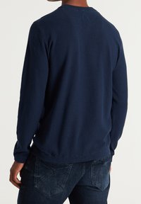 Pull texturé bleu marine à manches longues avec un col rond et des poignets côtelés, associé à un jean en denim foncé. La vue de dos montre un design simple.