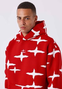 Sudadera roja con un gran patrón de estrellas blancas, que cuenta con un bolsillo frontal y un logo en el pecho. Textura suave, diseño oversized, capucha ajustable.
