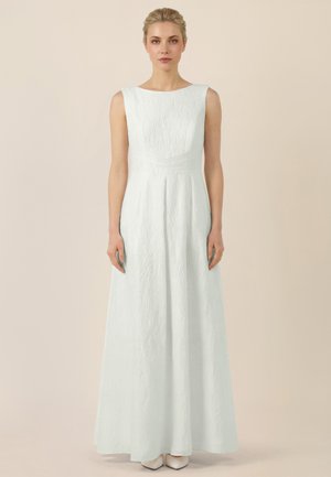 Frau steht und trägt ein ärmelloses, strukturiertes, weißes bodenlanges Kleid mit rundem Ausschnitt und weißen spitzen Schuhen.