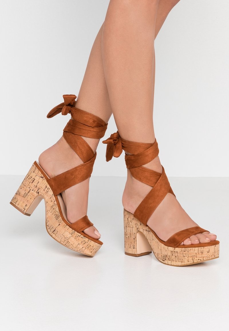 Brune ruskind hæle sandaler med kork-platforme, med krydsede ankelbånd og en bred tårem, glat tekstur og blokform.