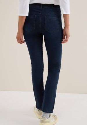 Persoon in donkerblauwe strakke jeans en witte sneakers, met de rug naar de camera, tegen een effen achtergrond.