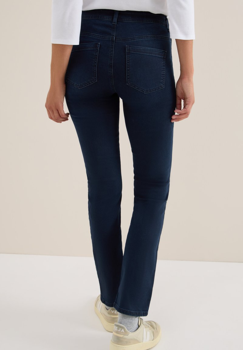 Persoon in donkerblauwe strakke jeans en witte sneakers, met de rug naar de camera, tegen een effen achtergrond.