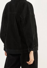 Chaqueta de denim negra con un corte relajado, que presenta un collar clásico, costuras visibles y puños enrollados. La tela tiene una textura sutil.