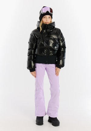 Schwarze Daunenjacke mit glänzender Oberfläche, tailliert, kombiniert mit lavendelfarbenen Schneehosen und schwarzen Winterstiefeln. Die Accessoires umfassen eine Mütze und eine Skibrille.