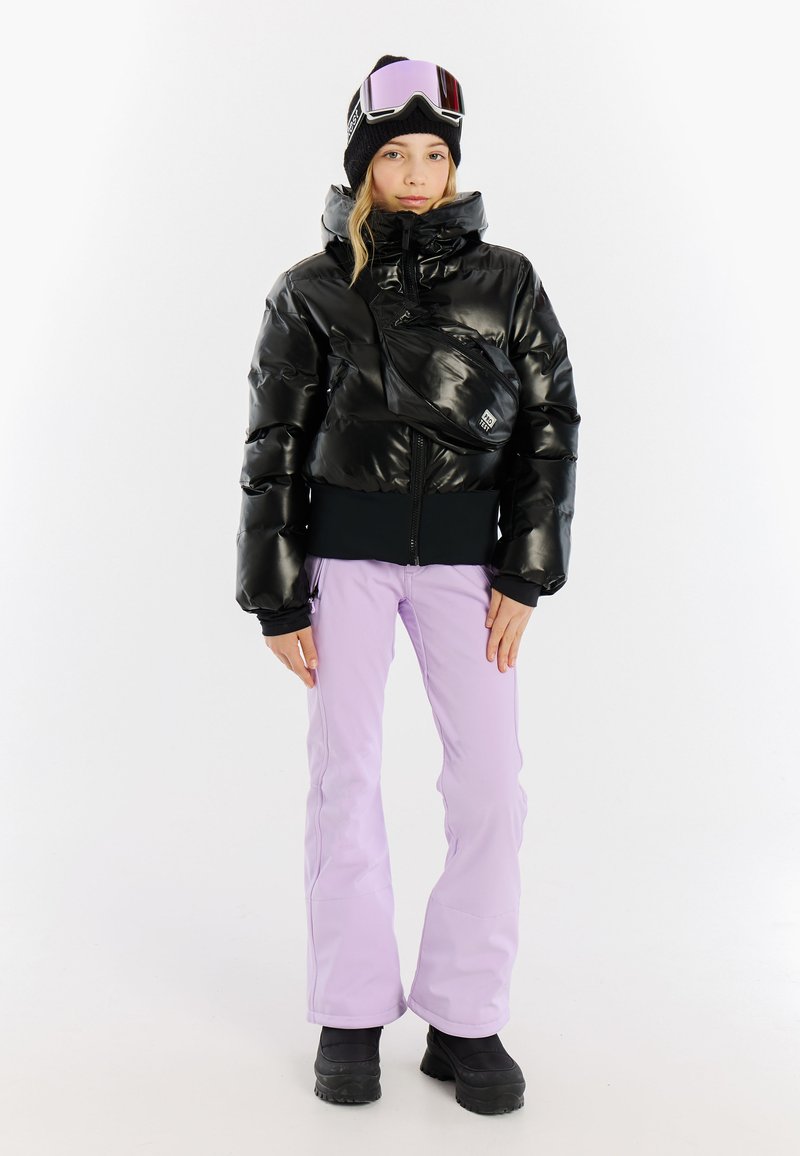 Zwarte pufferjack met een glanzende afwerking, getailleerde taille, gecombineerd met lavendelkleurige sneeuwbroeken en zwarte winterschoenen. Accessoires omvatten een beanie en een skibril.