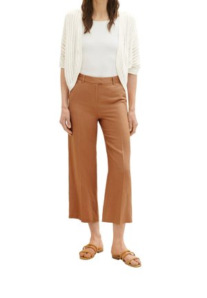 Donna che indossa un cardigan lavorato a maglia bianco a maniche corte, canottiera bianca, pantaloni larghi marroni e sandali bassi color cuoio, in piedi immobile.