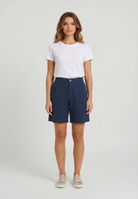 Mujer de pie con una camiseta blanca de manga corta, shorts de tiro alto azul marino con botón y zapatillas beige sobre un fondo liso.