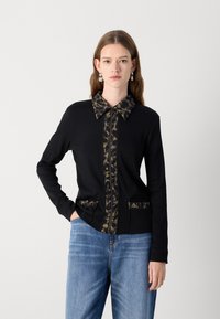Zwarte cardigan met een gemusterde kraag en zakken, met een getailleerd ontwerp. De kraag en zakken hebben een camouflageprint.