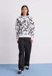 Person with long hair wearing a black-and-white abstract patterned sweater, loose black pants, and black sneakers, standing on a blue floor.